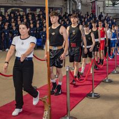 boxers wearing all black, all blue, or all red kit, walking down a red carpet in gym, following woman in black trousers and white top. 