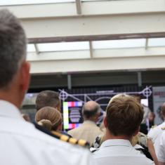 back of heads of naval personnel facing stage where there is a person speaking