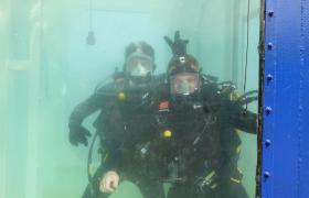 Two men in a water tank in wet suits, one is doing bunny ears with his fingers above his friends head.