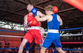 Two boxers in a ring during a match, one in red and the other in blue, exchanging punches under bright lights with a referee and audience in the background.