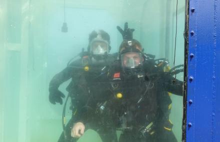 Two men in a water tank in wet suits, one is doing bunny ears with his fingers above his friends head.