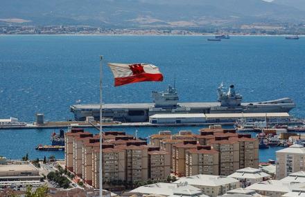 View of Gibraltar with the country's flag waving above a historic stone fortress, overlooking the Mediterranean Sea with naval ships and mountains in the background.
