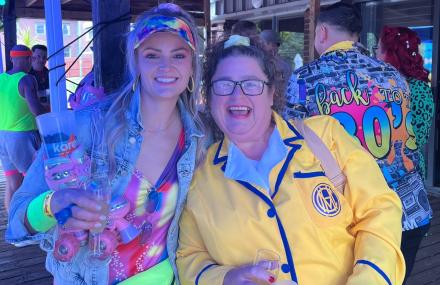 Two people smiling at a colorful event, one holding a clear cup, wearing vibrant and playful attire, including a jacket with "Back to the 80s" text.