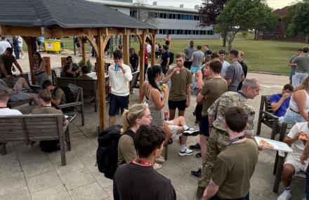 Group of people in casual and military attire enjoying an outdoor gathering with food, seated and standing in a courtyard area with benches and a wooden gazebo.