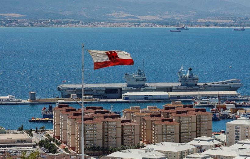 View of Gibraltar with the country's flag waving above a historic stone fortress, overlooking the Mediterranean Sea with naval ships and mountains in the background.