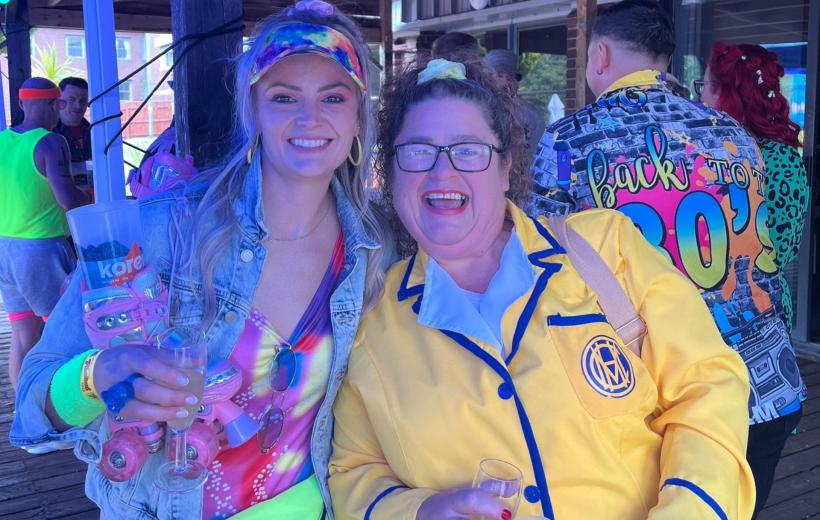 Two people smiling at a colorful event, one holding a clear cup, wearing vibrant and playful attire, including a jacket with "Back to the 80s" text.