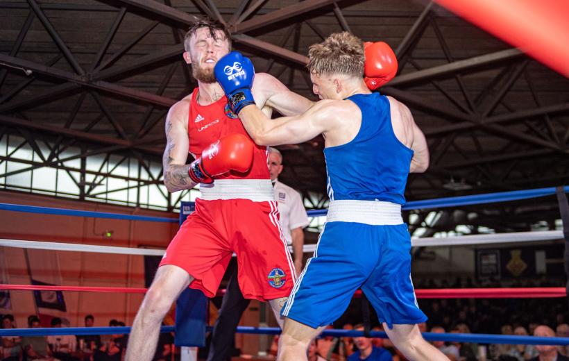 Two boxers in a ring during a match, one in red and the other in blue, exchanging punches under bright lights with a referee and audience in the background.