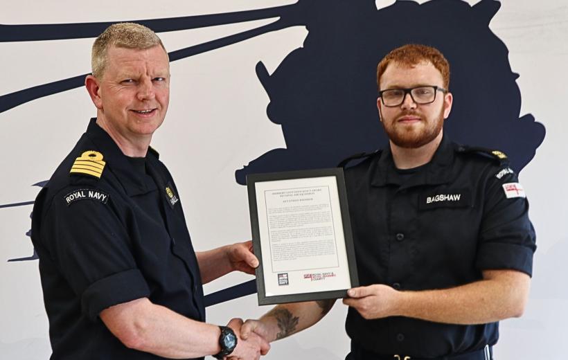 Royal Navy personnel shaking hands as one presents the other with a framed Herbert Lott Efficiency Award. Both are in uniform, standing in front of a naval-themed backdrop.